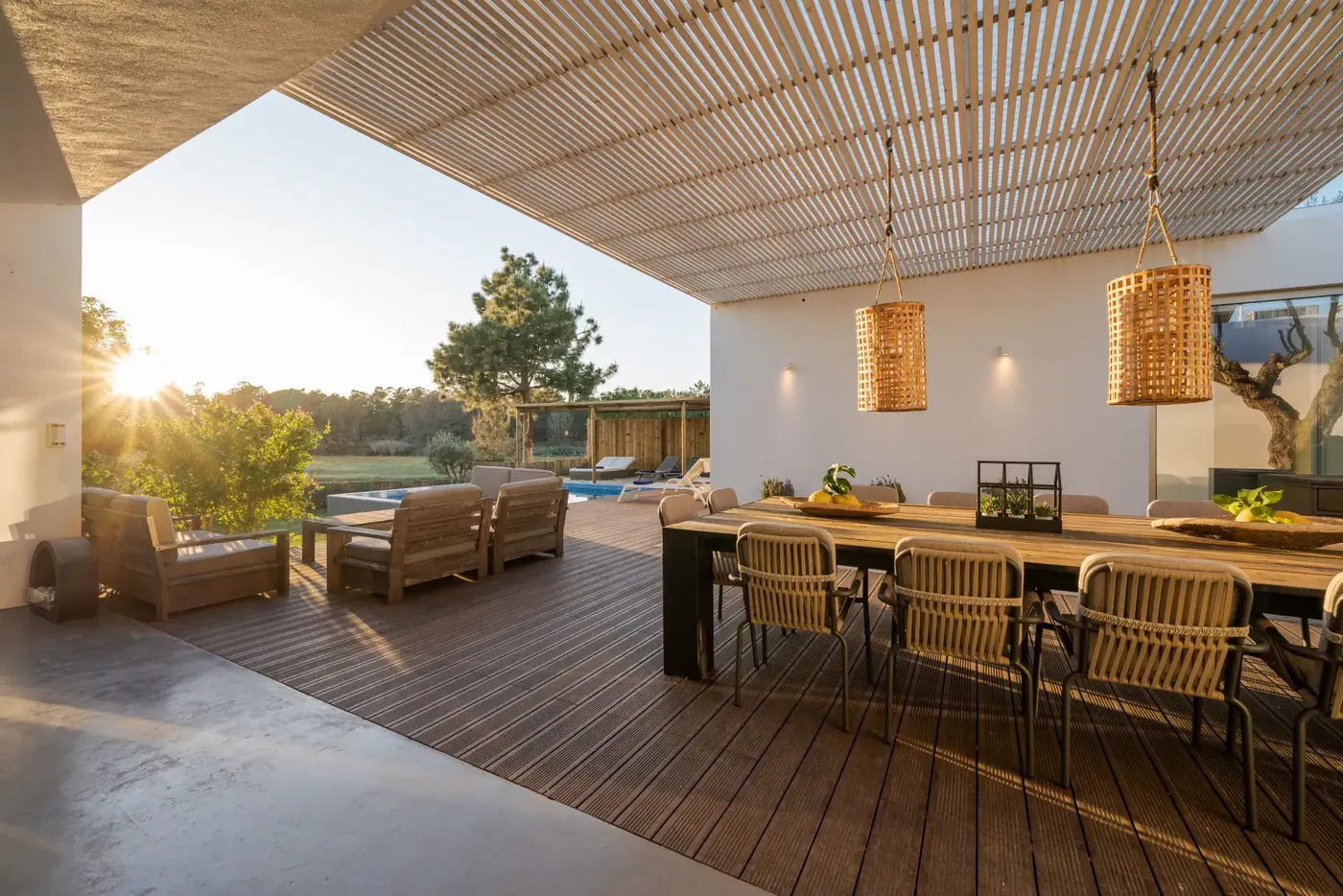 Covered outdoor terrace with dining table and lounge chairs beside a pool at sunset